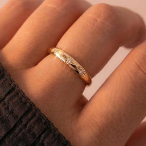 Peut inclure: Une bague dorée ornée de trois petites pierres précieuses transparentes. La bague a une surface lisse et brillante, reflétant la lumière. La bague est portée au doigt, avec une manche sombre en arrière-plan.