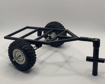 Può includere: Un rimorchio modello nero stampato in 3D con due grandi pneumatici fuoristrada neri. Il rimorchio ha un telaio rettangolare e un gancio di traino nero. I pneumatici hanno cerchi argentati e un battistrada robusto.