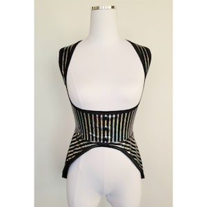 May include: A black and silver striped corset-style top with a halter neck design. The top is displayed on a white mannequin. The vertical stripes have a reflective, iridescent quality, creating a shimmering effect. The garment has a structured silhouette.