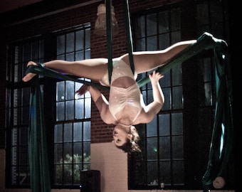 May include: A performer in a white sequined leotard and fishnet stockings is suspended upside down on green aerial silks. The performer is in a split position, with arms extended. The background includes windows and brick walls.