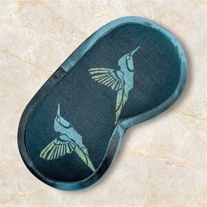 May include: A dark teal sleep mask with a satin border and a hummingbird design. The hummingbirds are light blue and green, with their wings spread. The mask is oval-shaped and designed to block out light.