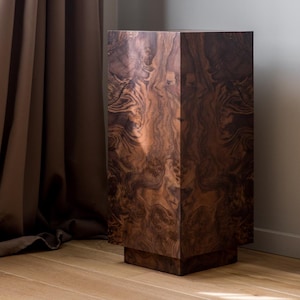 Op de afbeelding: Een hoge, rechthoekige houten sokkel met een rijk, donkerbruin wortelnotenpatroon. De sokkel heeft een vierkante basis en een bijpassende bovenkant, waardoor een opvallend display ontstaat. De houtnerf heeft wervelende patronen.