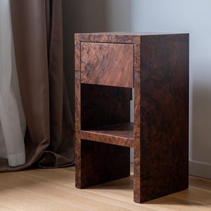 Può includere: Un comodino in legno marrone scuro con un cassetto e un ripiano aperto. La venatura del legno è visibile, creando un aspetto strutturato. Il comodino ha una forma rettangolare e si trova sul pavimento.