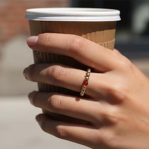 Könnte beinhalten: Ein goldener Perlenring mit einer einzelnen roten Perle wird an einem Finger getragen. Der Ring wird in einer Hand gehalten, die einen braunen Kaffeebecher aus Papier mit einem weißen Deckel hält. Der Hintergrund ist unscharf.