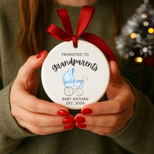 May include: A round white ceramic ornament featuring a red ribbon bow at the top. The text reads PROMOTED TO grandparents with a blue baby carriage illustration in the centre and the text BABY NATHAN DEC 2026 printed at the bottom.