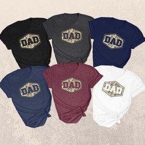 May include: Six short-sleeved t-shirts in various colors: black, charcoal, navy, burgundy, and white. Each shirt features a hexagon design with the word "DAD" in a bold font, set against a camouflage pattern.