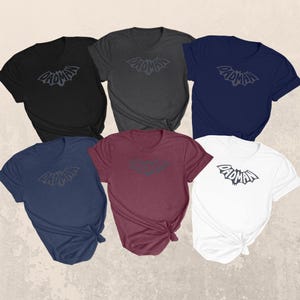 May include: A collection of six t-shirts in various colors: black, charcoal, navy, burgundy, and white. Each shirt features a gray bat-shaped logo with the word "DADMAN" in a stylized font. The shirts are neatly arranged, showcasing the design.