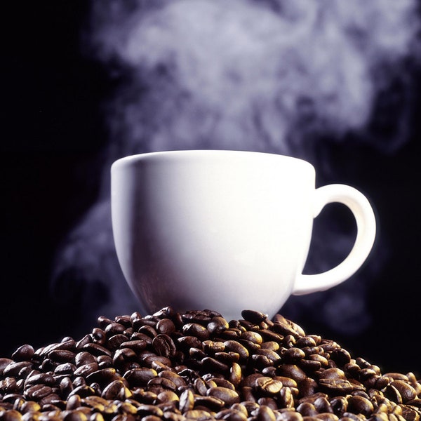 May include: A white ceramic coffee mug with steam rising from it, sitting on a bed of roasted coffee beans.