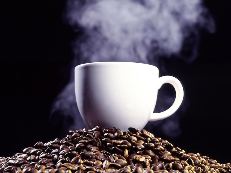 May include: A white ceramic coffee mug with steam rising from it, sitting on a bed of roasted coffee beans.