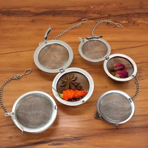 May include: Five silver tea infusers with chains. The infusers are open and show different ingredients inside, including star anise, peppercorns, and dried rosebuds.