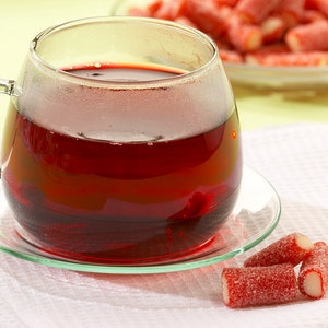 Puede incluir: Una taza de vidrio llena de t&eacute; rojo se encuentra sobre un platillo blanco con caramelos rojos y blancos sobre una superficie blanca.