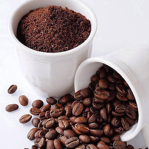 May include: Two white ceramic mugs, one filled with ground coffee and the other with whole coffee beans. The beans are spilling out of the mug onto a white surface.