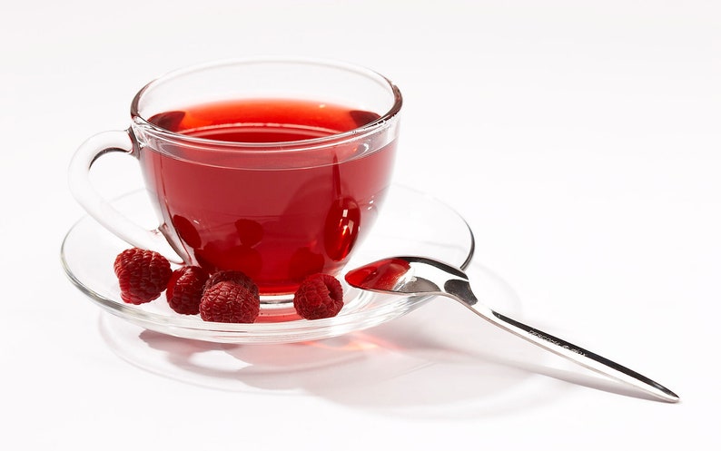 Peut inclure: Une tasse &agrave; th&eacute; en verre transparent remplie de th&eacute; rouge &agrave; la framboise, pos&eacute;e sur une soucoupe avec quatre framboises fra&icirc;ches. Une cuill&egrave;re &agrave; caf&eacute; en argent repose sur la soucoupe.
