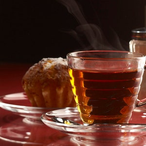 Könnte beinhalten: Nahaufnahme einer gläsernen Teetasse gefüllt mit dunklem Tee, die auf einem Untersetzer mit einem kleinen, runden Gebäck daneben steht. Im Hintergrund befindet sich ein gläserner Zuckerstreuer mit weißem Zucker.