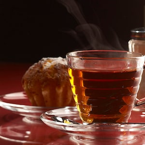 Puede incluir: Primer plano de una taza de t&eacute; de vidrio llena de t&eacute; oscuro, sentada sobre un platillo con un peque&ntilde;o pastel redondo. La taza de t&eacute; est&aacute; sobre una superficie roja con un dispensador de az&uacute;car en el fondo.