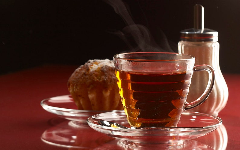 Puede incluir: Primer plano de una taza de t&eacute; de vidrio llena de t&eacute; oscuro, sentada sobre un platillo con un peque&ntilde;o pastel redondo. La taza de t&eacute; est&aacute; sobre una superficie roja con un dispensador de az&uacute;car en el fondo.