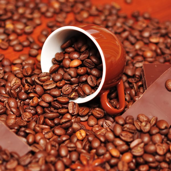 May include: A brown ceramic coffee cup filled with roasted coffee beans, spilling onto a surface covered in coffee beans. Two pieces of dark chocolate are also on the surface.