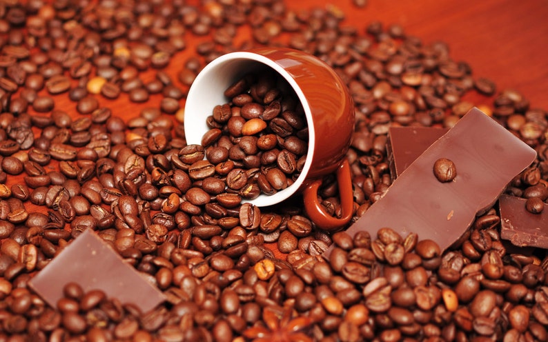 May include: A brown ceramic coffee cup filled with roasted coffee beans, spilling onto a surface covered in coffee beans. Two pieces of dark chocolate are also on the surface.