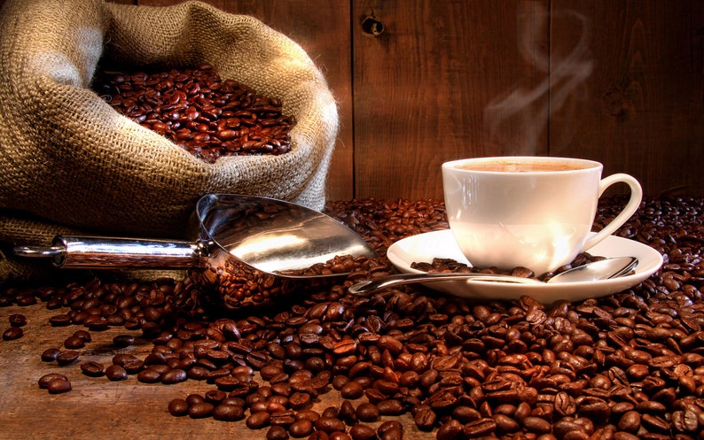 May include: A white coffee cup with steam rising from it sits on a saucer with a spoon. The cup is surrounded by coffee beans, and a burlap sack filled with coffee beans is in the background.