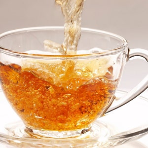 Könnte beinhalten: Eine klare Teetasse und Untertasse aus Glas werden mit bernsteinfarbenem Tee gefüllt, während mehr Tee hineingegossen wird. Der Tee wirbelt und spritzt, wodurch Blasen und Bewegung entstehen. Der Hintergrund ist eine weiche, neutrale Farbe.