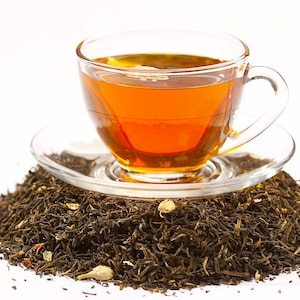 Peut inclure: Une tasse en verre remplie de th&eacute; ambr&eacute; repose sur une soucoupe au-dessus d'un tas de feuilles de th&eacute; en vrac. Les feuilles de th&eacute; sont un m&eacute;lange de brun et de vert, avec quelques p&eacute;tales de fleurs blanches.