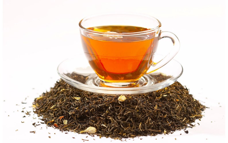 Peut inclure: Une tasse en verre remplie de th&eacute; ambr&eacute; repose sur une soucoupe au-dessus d'un tas de feuilles de th&eacute; en vrac. Les feuilles de th&eacute; sont un m&eacute;lange de brun et de vert, avec quelques p&eacute;tales de fleurs blanches.
