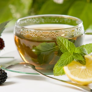 Peut inclure: Une tasse de th&eacute; vert dans un verre avec des feuilles de menthe fra&icirc;che, un quartier de citron et des m&ucirc;res sur une surface blanche.