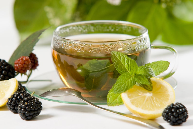 Peut inclure: Une tasse de th&eacute; vert dans un verre avec des feuilles de menthe fra&icirc;che, un quartier de citron et des m&ucirc;res sur une surface blanche.