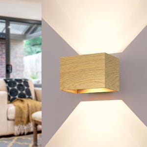 Può includere: Applique da parete rettangolare moderna con finitura in legno di rovere chiaro. L'apparecchio è montato su una parete grigia, con luce che emana dall'alto e dal basso, creando un effetto di illuminazione unico. L'interno dell'applique si illumina.