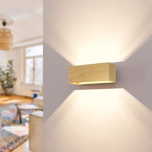 Può includere: Applique da parete rettangolare in legno con finitura a venatura naturale. L'applique emette luce verso l'alto e verso il basso, creando un bagliore soffuso sulla parete. L'apparecchio è montato su una parete grigio chiaro.