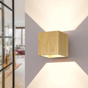 Op de afbeelding: Vierkante wandlamp van licht hout. De lamp is gemonteerd op een grijze muur, met licht dat van boven en beneden komt. Het ontwerp is modern en minimalistisch.