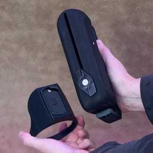 Puede incluir: Dos dispositivos electrónicos negros se sostienen frente a un fondo marrón. Uno es largo y rectangular con un botón y una ranura. El otro es un anillo negro con una base rectangular.