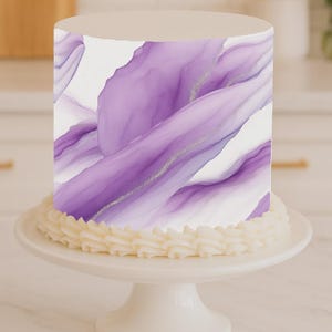 May include: A round cake with a white base and a purple and white abstract design. The cake sits on a white cake stand with a decorative cream-colored frosting border.