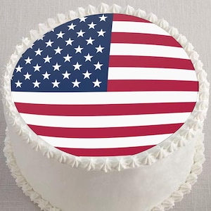 May include: A round white cake decorated with a frosting border. The cake is topped with an edible image of the American flag, featuring red and white stripes and a blue field with white stars. A festive dessert.