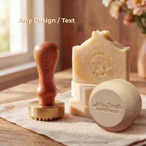 Könnte beinhalten: Ein Seifenstempel aus Messing mit Holzgriff neben einem Stapel rechteckiger Seifenstücke und einem runden Seifenstück. Der Stempel zeigt ein Bienen- und Honiglöffel-Motiv. Die Seifen sind mit Handmade Soap und John Smith Handmade geprägt.