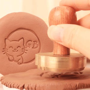 May include: A wooden handled stamp with a brass base is pressed into clay, creating an impression of a smiling cat and the letters "GB". The clay is a light brown color. The stamp is being held by a person's hand.