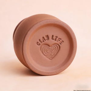 May include: A terracotta-coloured ceramic vessel base featuring an embossed stamp that reads CLAY LIFE above a heart shape filled with a fingerprint-like swirl pattern. The circular bottom is recessed within a smooth, rounded outer rim.