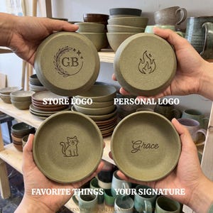 May include: Four round, sage green ceramic dishes with engraved designs. One features the letters GB inside a laurel wreath, one a flame icon, one a sitting cat, and one the name Grace in script. The dishes have a matte, textured finish.