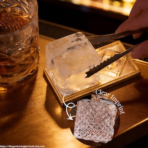 May include: A clear ice cube held by metal tongs, resting on a small gold tray. A glass of amber liquid is in the background. The image includes a smaller inset image of a patterned glass with the text "Clear Imprint".