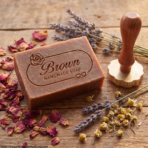 Può includere: Una saponetta artigianale marrone con la scritta "Brown" incisa, insieme a una rosa e due cuori. Il sapone è circondato da fiori secchi, lavanda e un timbro di legno.