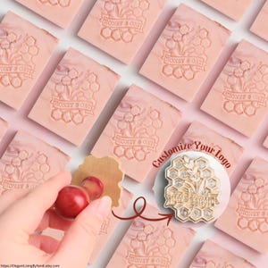 May include: Pink rectangular soaps with a honeycomb and floral design, stamped with "HONEY & OAT". A wooden stamp with a red handle is shown next to the soaps. The text "Customize Your Logo" is also visible.