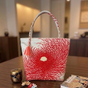 以下が含まれることがあります： クリーム色のトートバッグで、前面に鮮やかな赤色の菊の刺繍が施されています。バッグには編み込みの持ち手があり、小さな赤いタッセルが付いています。刺繍は細い放射状の線で詳細に描かれています。