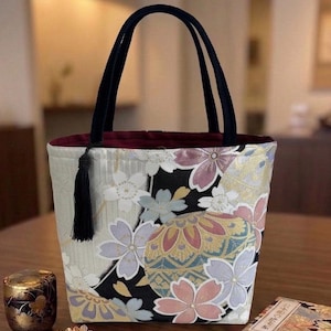 以下が含まれることがあります： 黒、クリーム、ゴールドの桜と円形モチーフの花柄トートバッグ。黒いハンドルが2本付いており、内側は濃い赤色の裏地で、開口部の片側には黒いタッセルがぶら下がっています。