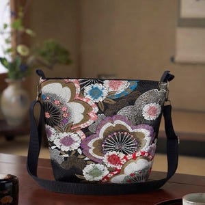 以下が含まれることがあります： 白、ピンク、紫、青の花々が描かれた鮮やかなマルチカラーのフローラル柄の黒いショルダーバッグ。バッグは構造化された先細りの形状で、金属製のリングに取り付けられた調節可能な黒い布製のストラップが付いています。