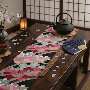 May include: A dark wooden table is set with a black teapot, a blue fan, and a floral runner. A vase of flowers, a cup of tea, and a small basket are also on the table. The runner features pink, red, and blue floral patterns.