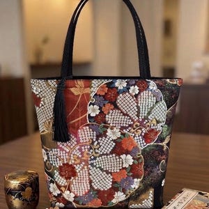 以下が含まれることがあります： 赤、金、白、紫の色合いで、鮮やかな多色の花柄と幾何学模様が施された暗い色の生地のトートバッグ。バッグには2本の黒い編み込みハンドルがあり、上部の開口部の片側には黒いタッセルがぶら下がっています。