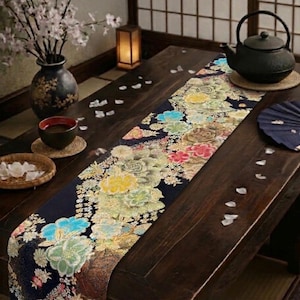 May include: A dark wooden table is adorned with a floral runner featuring colorful flowers. A black teapot, a cup, and a vase with white flowers are also on the table. The scene is set in a room with traditional Japanese decor.