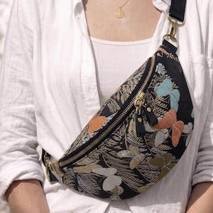 以下が含まれることがあります： 水色、ピーチ、ゴールド、シルバーの色合いで蝶とシダの葉の複雑な織り模様が施された黒のクロスボディバッグ。ゴールドトーンの金属製ジッパーには、2つの黒いレザーの引き手が付いており、黒いストラップが付属しています。