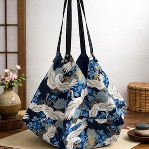 Puede incluir: Bolso tote azul marino con un estampado japonés de grullas y flores. El bolso tiene asas negras y una forma geométrica. El diseño presenta grullas blancas, flores azules y detalles dorados.