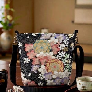 以下が含まれることがあります： ピンク、黄色、紫、白の色合いで、密集した複雑な花柄が施された黒いショルダーバッグ。バッグは長方形で、金属製の金具で取り付けられた黒い調節可能なストラップがあり、背景には質感のある暗い生地が使用されています。